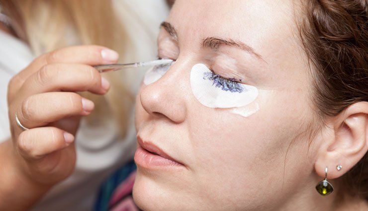 Person getting eyelash extensions, under-eye pads in place. Technician uses tweezers, close-up.