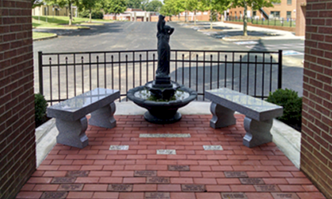A memorial garden courtyard featuring a central water fountain, two stone benches, and a brick pathway with engraved pavers.