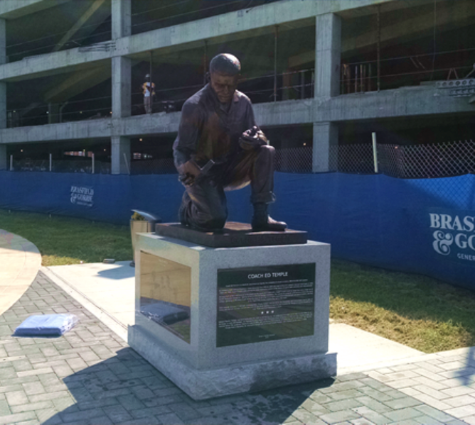 A bronze statue of a person kneeling and working on a small object, mounted on a granite pedestal in an outdoor setting.