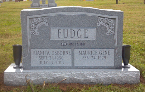 A gray granite double headstone for the Fudge family, engraved with names and dates, standing in a grassy cemetery.