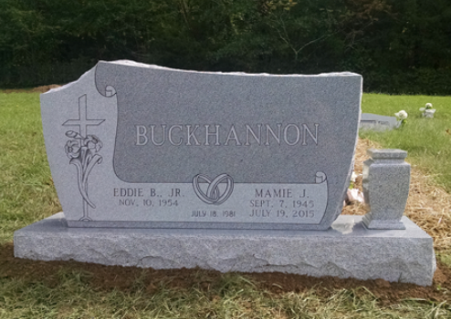 A gray granite headstone for the Buckhannon family, featuring a cross, roses, and wedding rings, set in a grassy field.