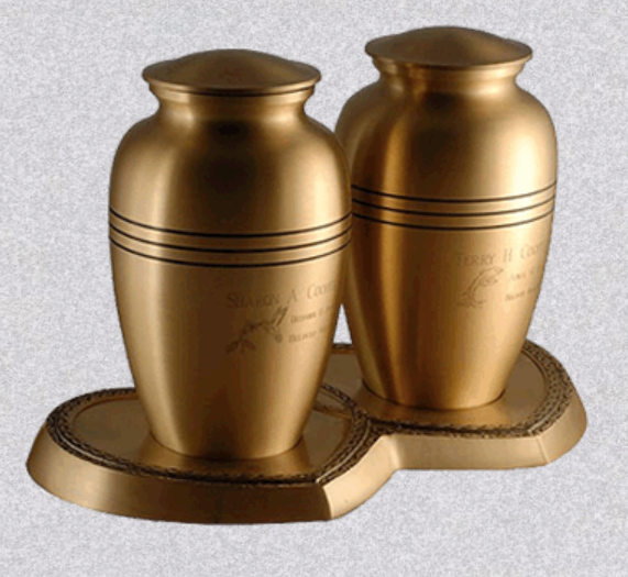 Two gold-toned metallic funeral urns displayed together on a shared heart-shaped base.