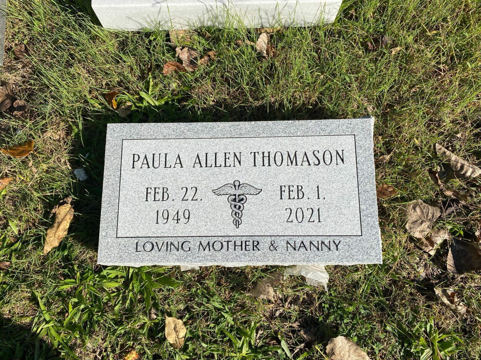 A gray stone grave marker for Paula Allen Thomason (1949–2021) with a caduceus symbol and the words Loving Mother & Nanny.