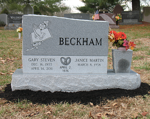 A gray granite headstone for Gary and Janice Beckham, featuring an engraving of praying hands and a vase of flowers.