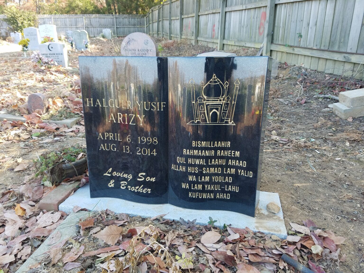A black, book-shaped headstone with gold text and a mosque engraving, set in a cemetery surrounded by dry leaves.