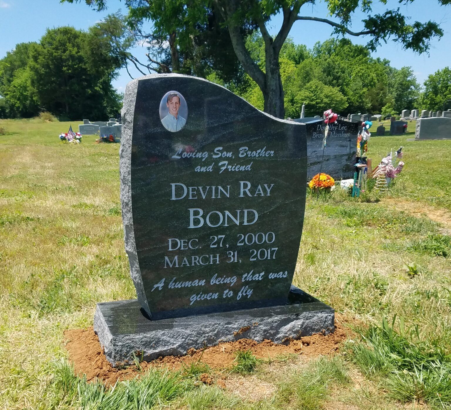 A black granite headstone with a portrait and inscription for Devin Ray Bond, set in a green, sunny cemetery.