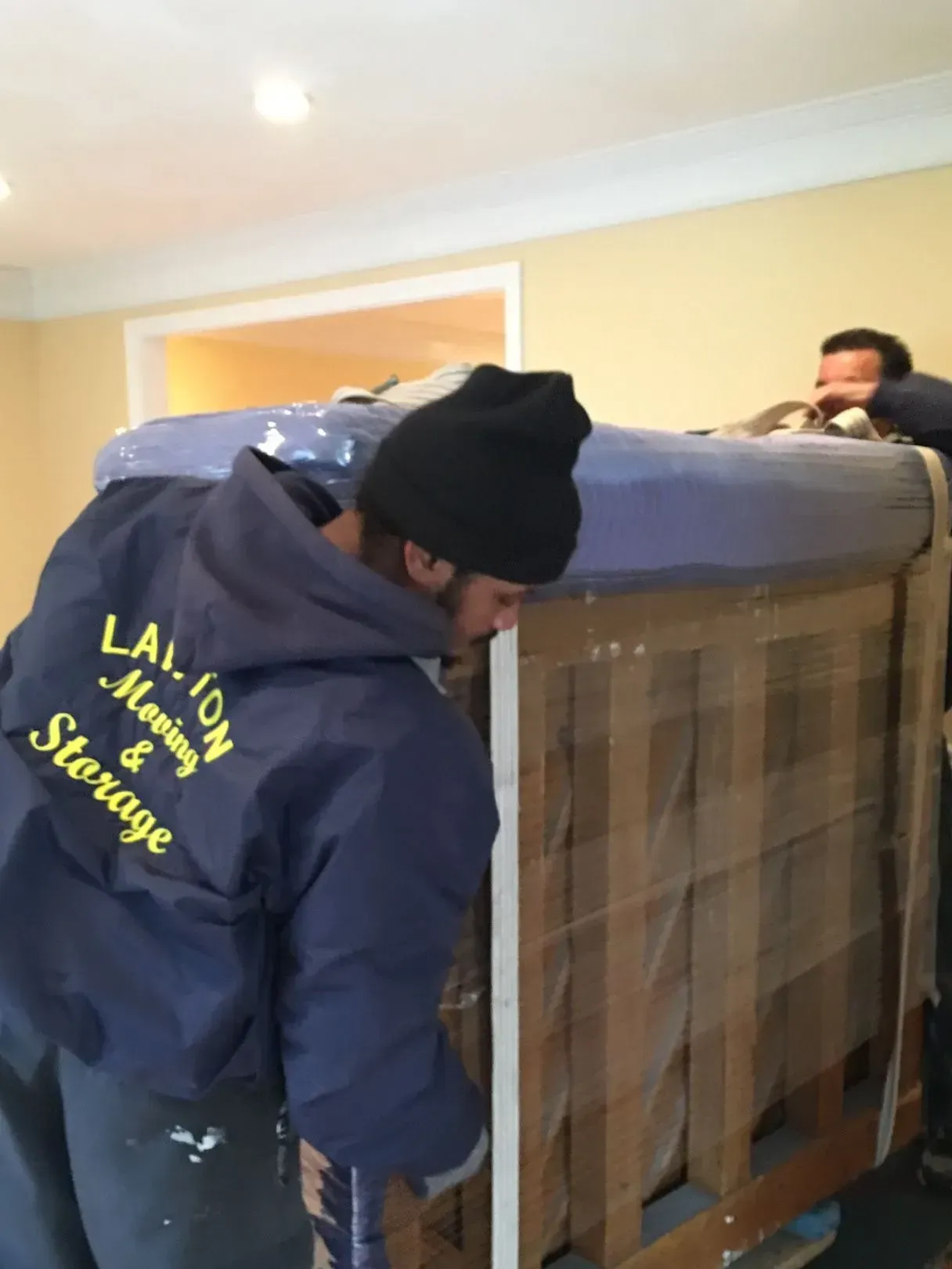 Two movers in blue jackets load a large, packaged item indoors.