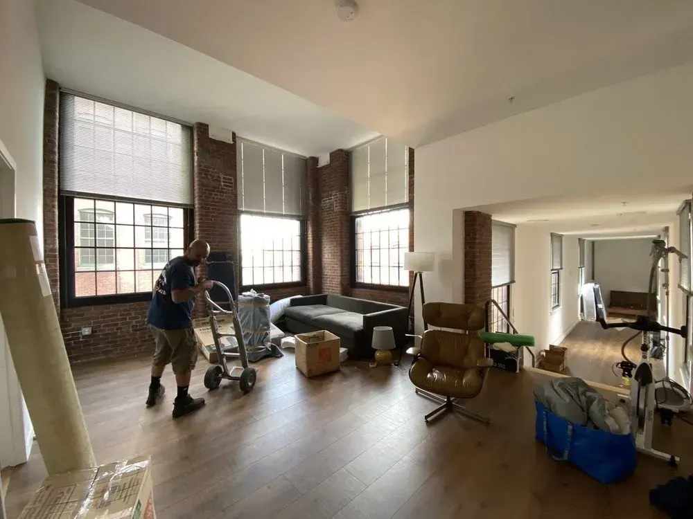 Man moves boxes on a dolly into a large, empty room with brick walls and large windows.