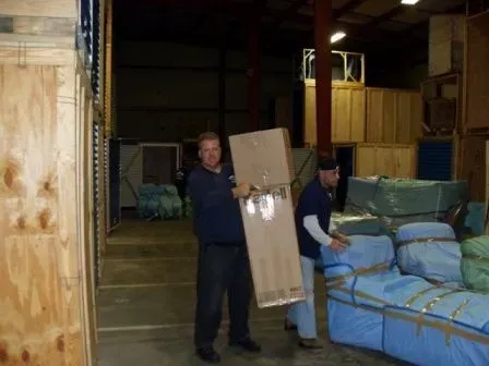 Two men moving a large cardboard box in a warehouse. One man holds the box, the other assists; surrounded by crates and furniture.