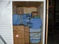 Storage unit filled with boxes and blue-wrapped items. White door, wooden walls.