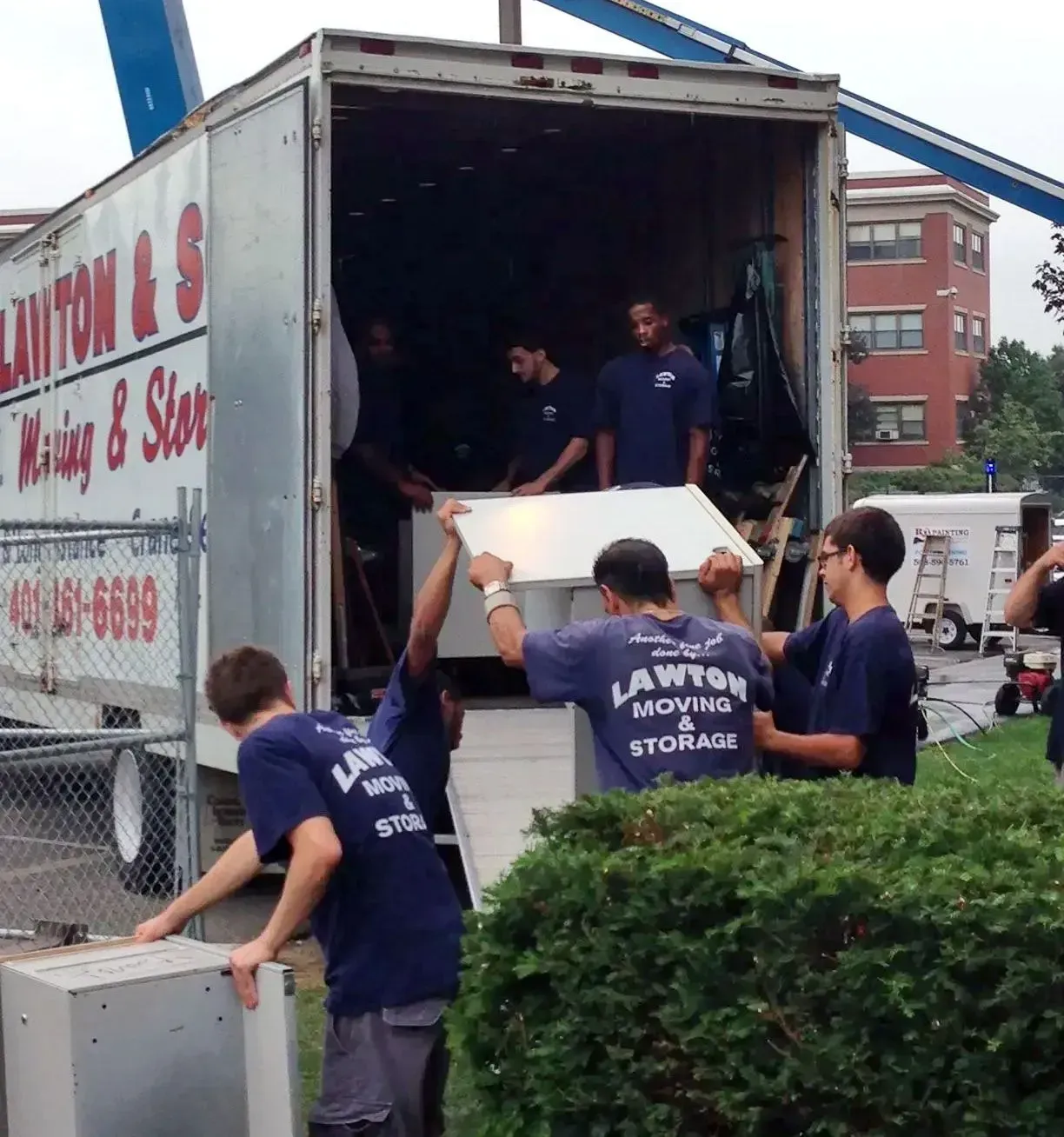 Movers in blue shirts unload furniture from a truck, with a building and a fence in the background.
