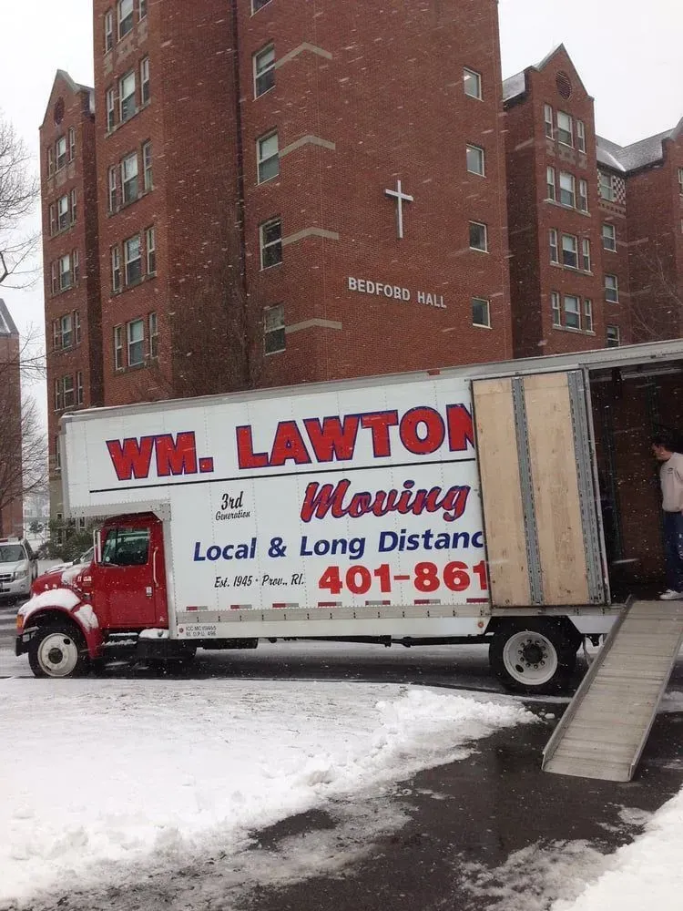 A red and white moving truck from WM. Lawton Moving parked in front of a brick building, on a snowy day.