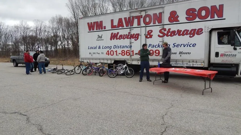 People gather around a moving truck with bicycles in front. A red table and moving van are visible in an outdoor setting.