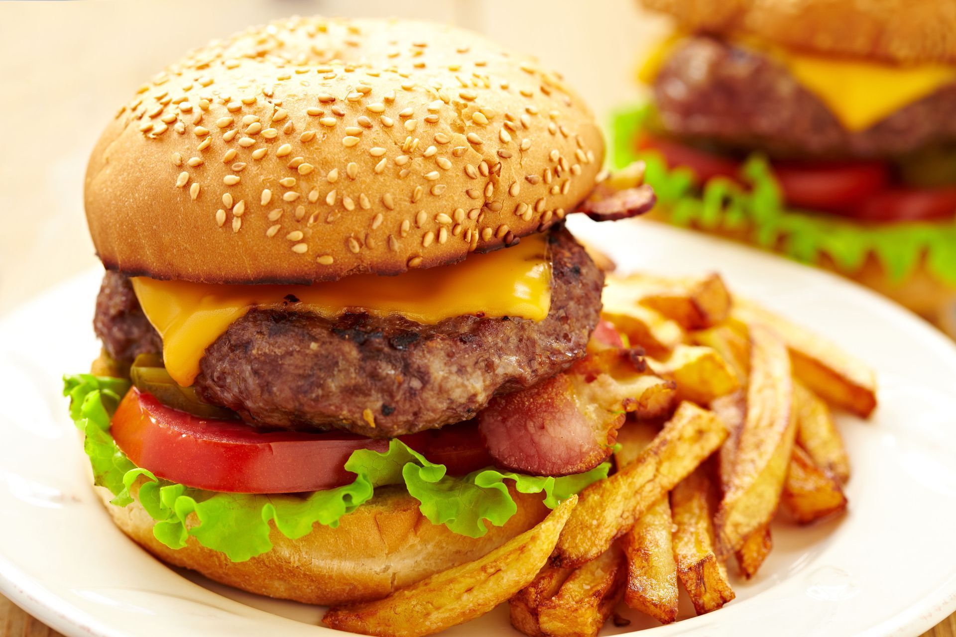 A cheeseburger with bacon, lettuce, and tomato on a plate with French fries, with another burger visible in the background.