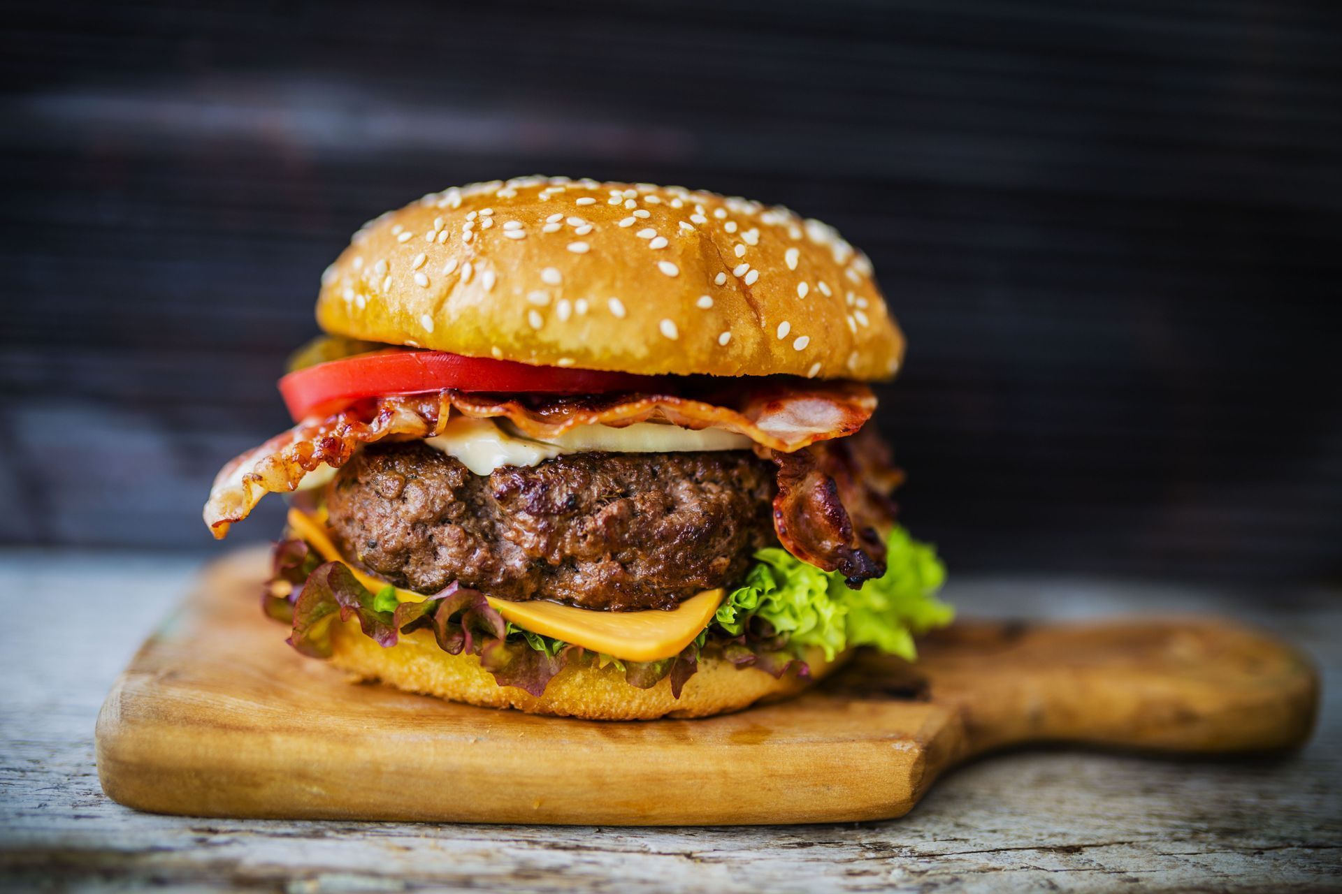 A bacon cheeseburger with lettuce, tomato, and cheese on a toasted sesame seed bun, served on a wooden cutting board.