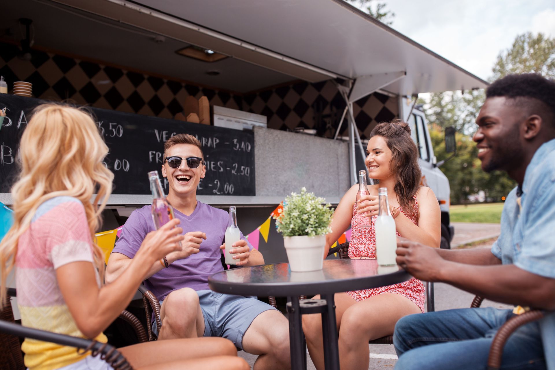 Four friends sitting at an outdoor table with drinks in front of a food truck, smiling and talking.