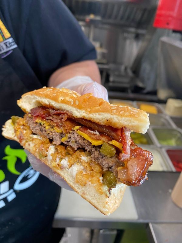 A close-up of a hand in a glove holding a cheeseburger with bacon and jalapeños in a kitchen.