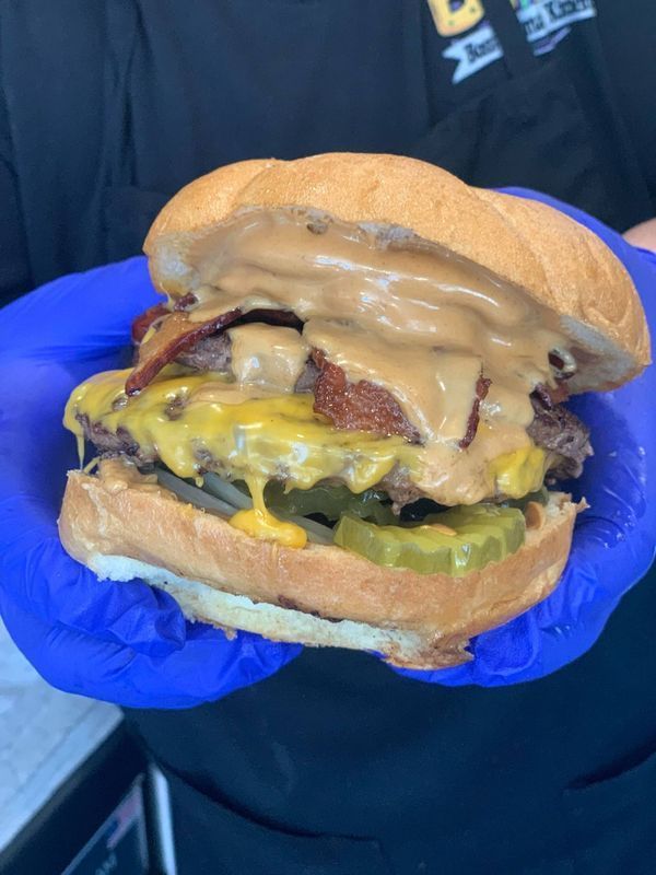 A person wearing blue gloves holds a double cheeseburger topped with peanut butter and bacon.