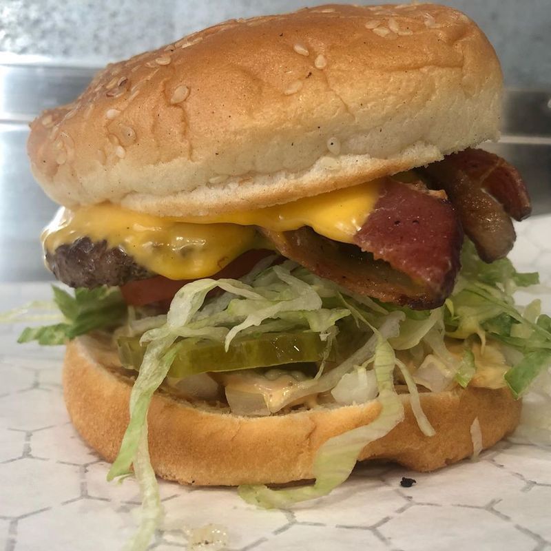 A cheeseburger topped with bacon, lettuce, and pickles on a toasted bun, sitting on white patterned paper.
