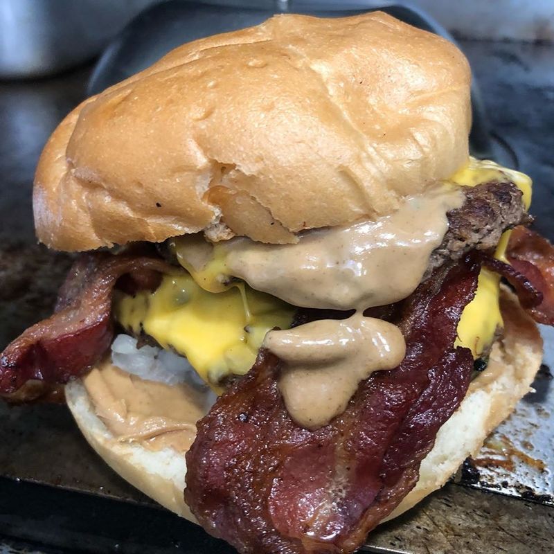 A double cheeseburger on a bun, topped with thick strips of crispy bacon and a creamy brown sauce on a flat-top grill.