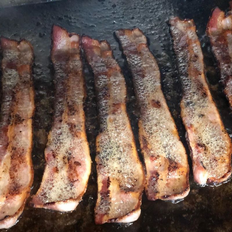 Six strips of bacon sizzling and seasoning on a hot, dark cooking surface.