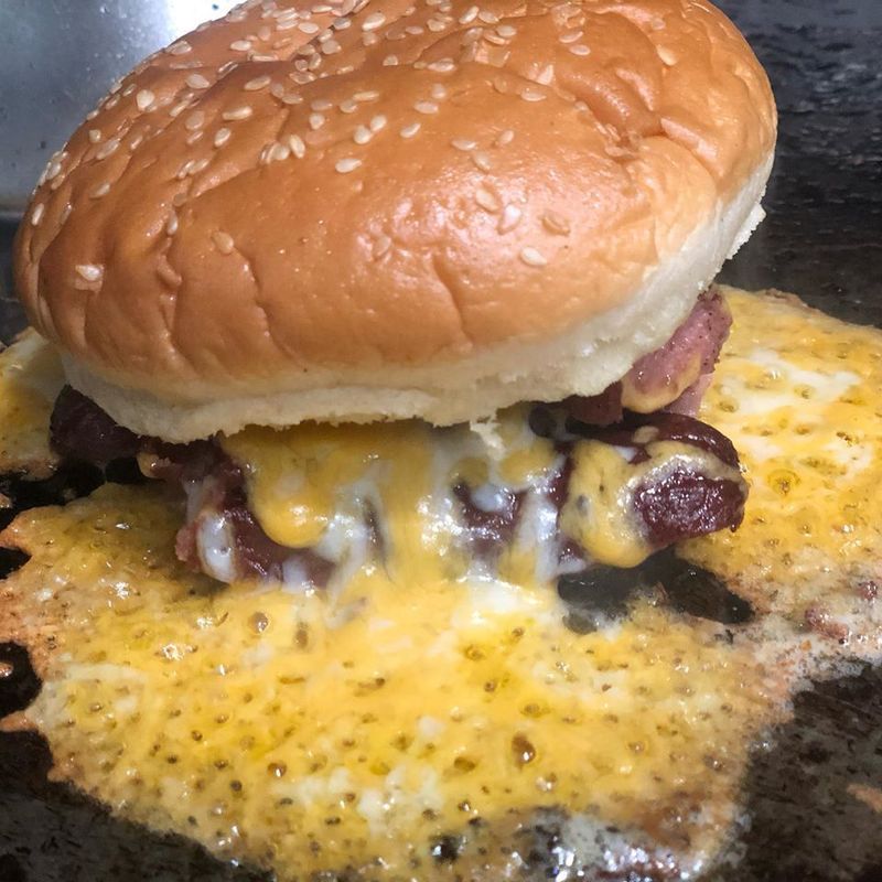 A cheeseburger cooking on a flat-top grill, featuring a sesame seed bun and a large, crispy melted cheese skirt.