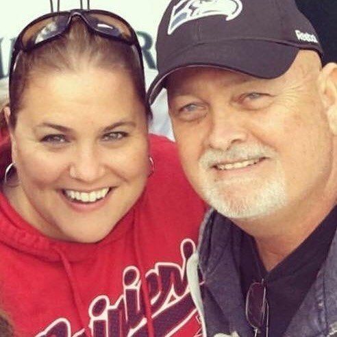 A smiling woman in a red hoodie and a man in a Seahawks baseball cap pose close together for a photo.