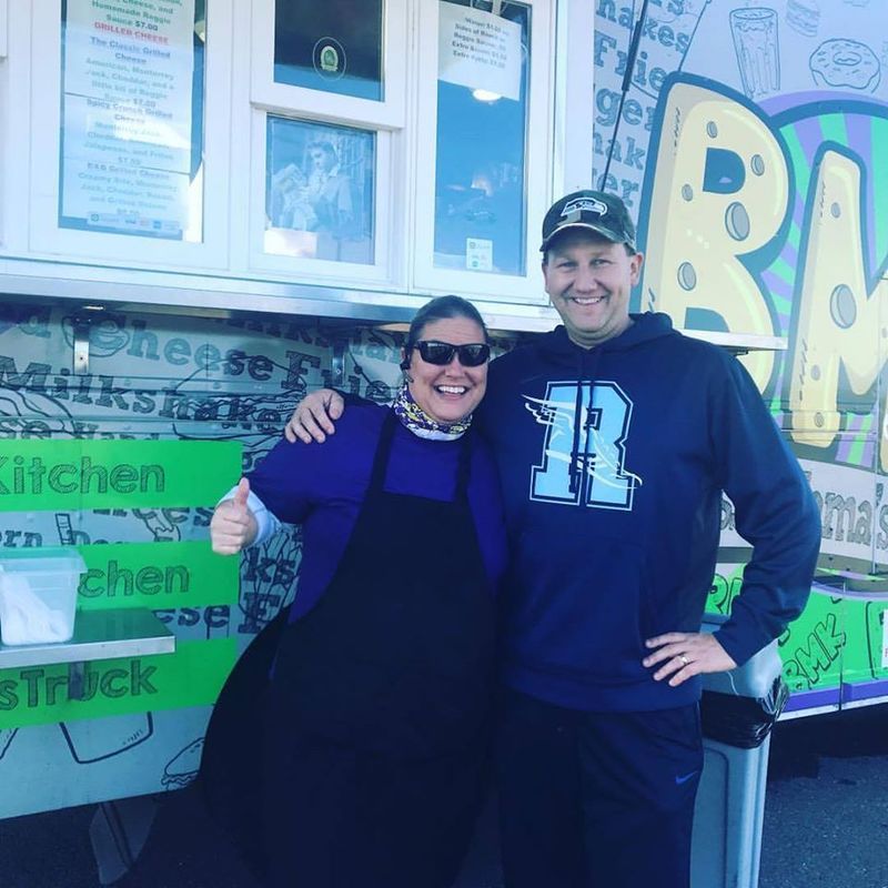 A smiling person in a dark apron and sunglasses standing next to a smiling person in a blue hoodie in front of a food truck.