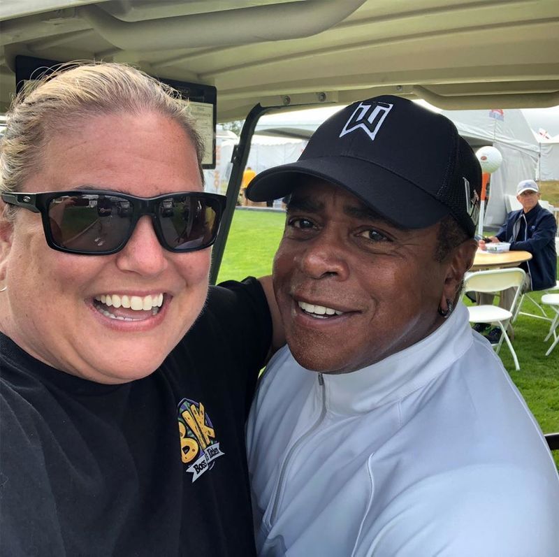 A smiling person takes a selfie with a person wearing a black cap inside a golf cart at an outdoor event.