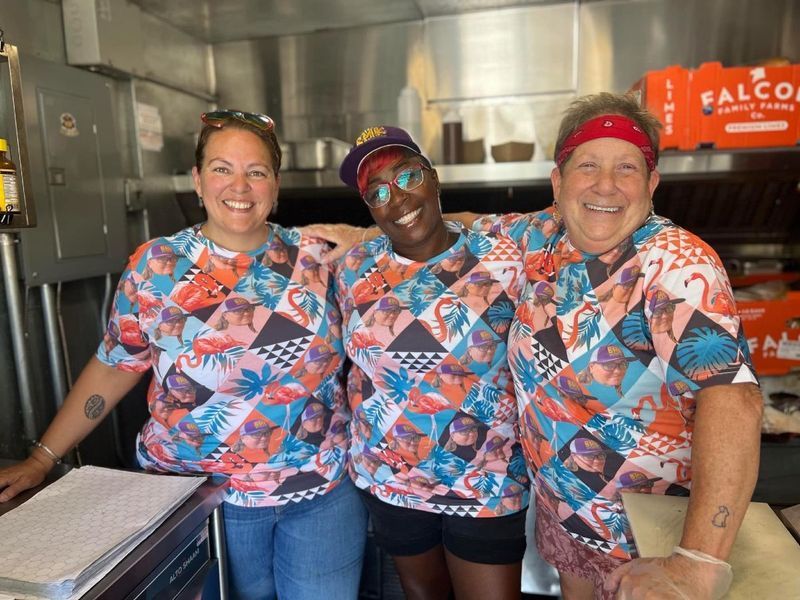 Three smiling people in matching, colorful patterned shirts stand behind a counter in a food truck workspace.