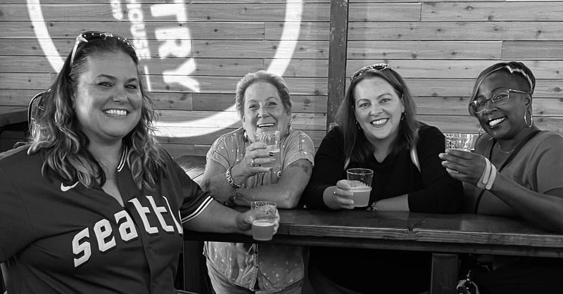 Four people smiling while holding glasses and sitting at a wooden table in front of a wall with a circular logo.