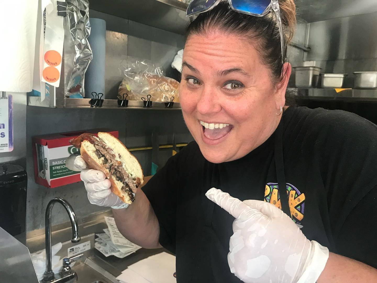 A smiling person wearing food service gloves holds a cheesesteak sandwich and points to it while in a kitchen.