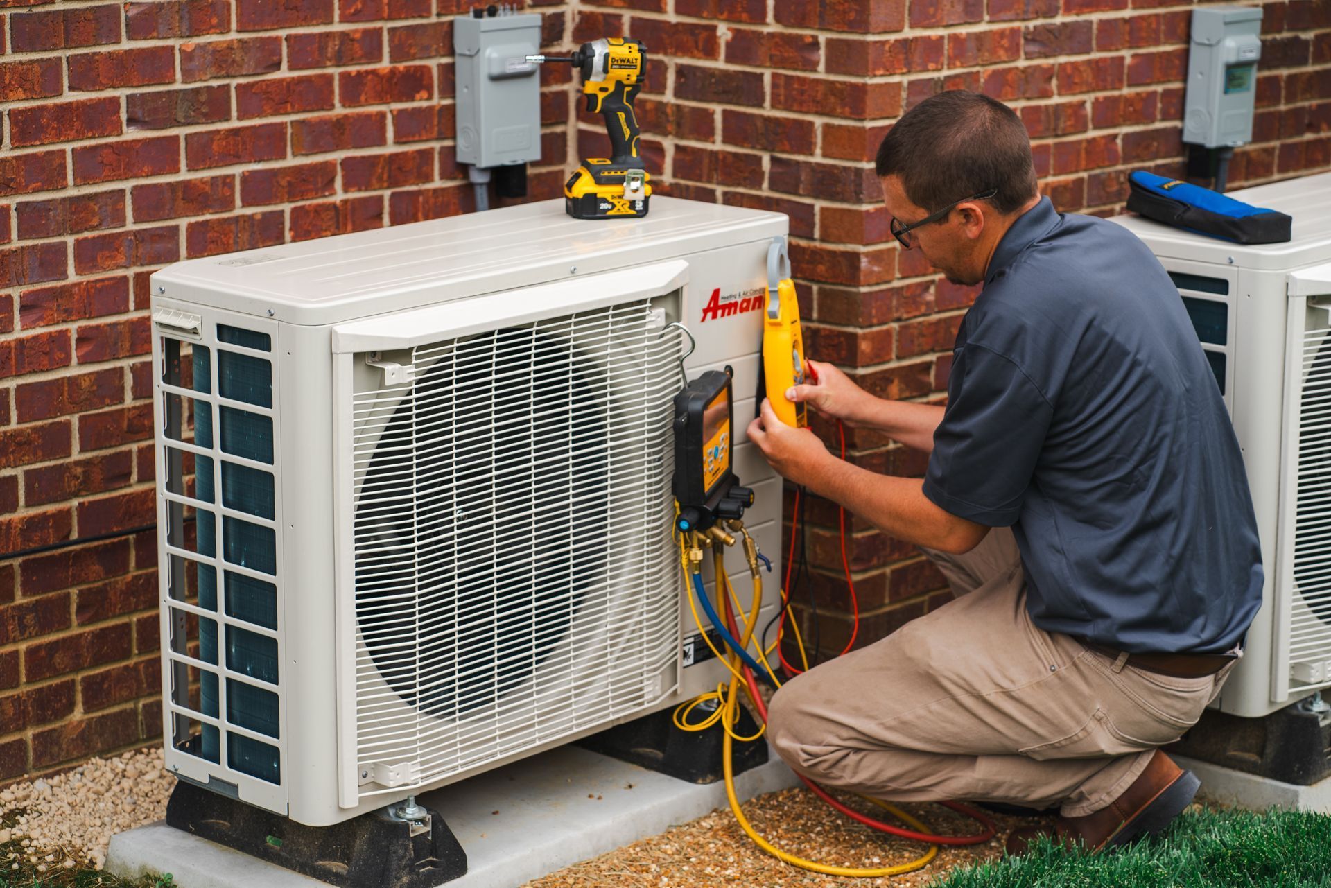 HVAC technician in tan pants, checking gauges on an outdoor air conditioning unit near a brick wall.