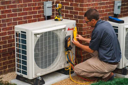 HVAC technician in tan pants, checking gauges on an outdoor air conditioning unit near a brick wall.