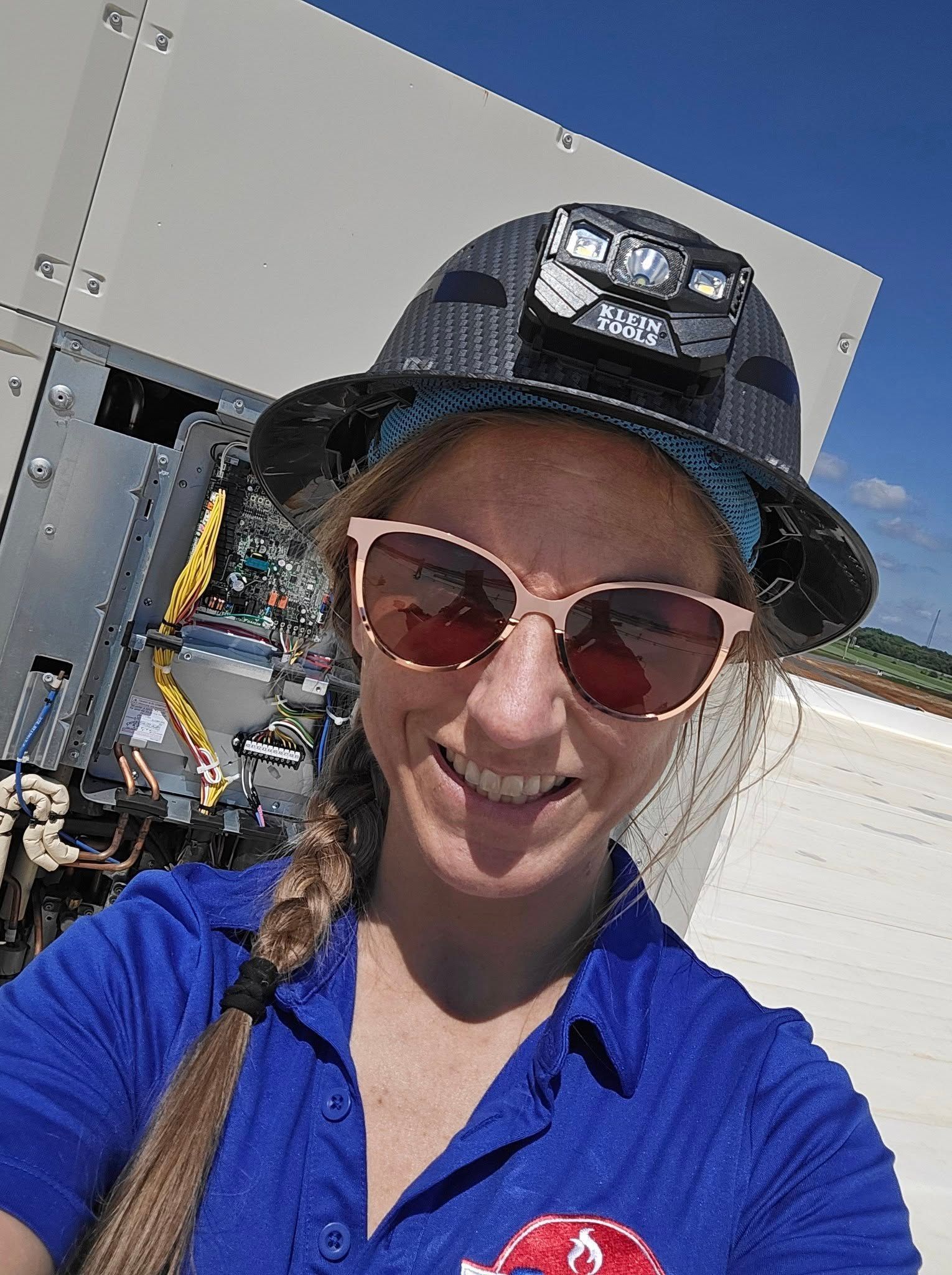 Woman in blue shirt and safety hat with sunglasses smiles at camera, outdoors.