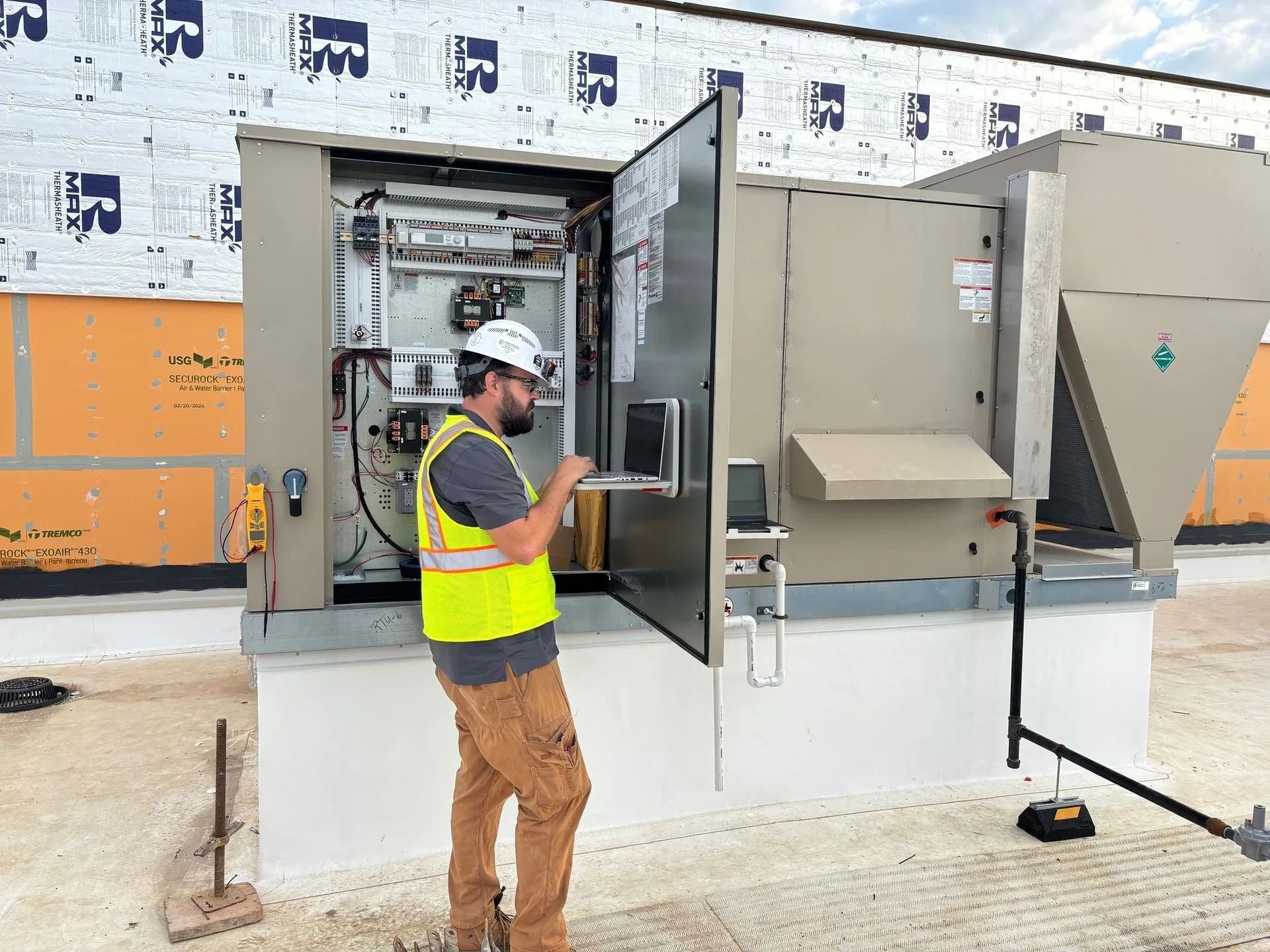HVAC technician in hard hat and vest, using a laptop to service rooftop air conditioning unit.