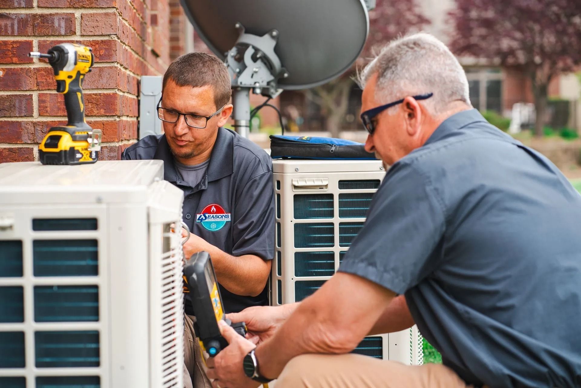 Two HVAC technicians inspecting an outdoor air conditioning unit. One holds a tool, the other a diagnostic device.