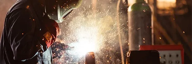 Person welding metal in a dim workshop, bright sparks flying amid tools and gas cylinders
