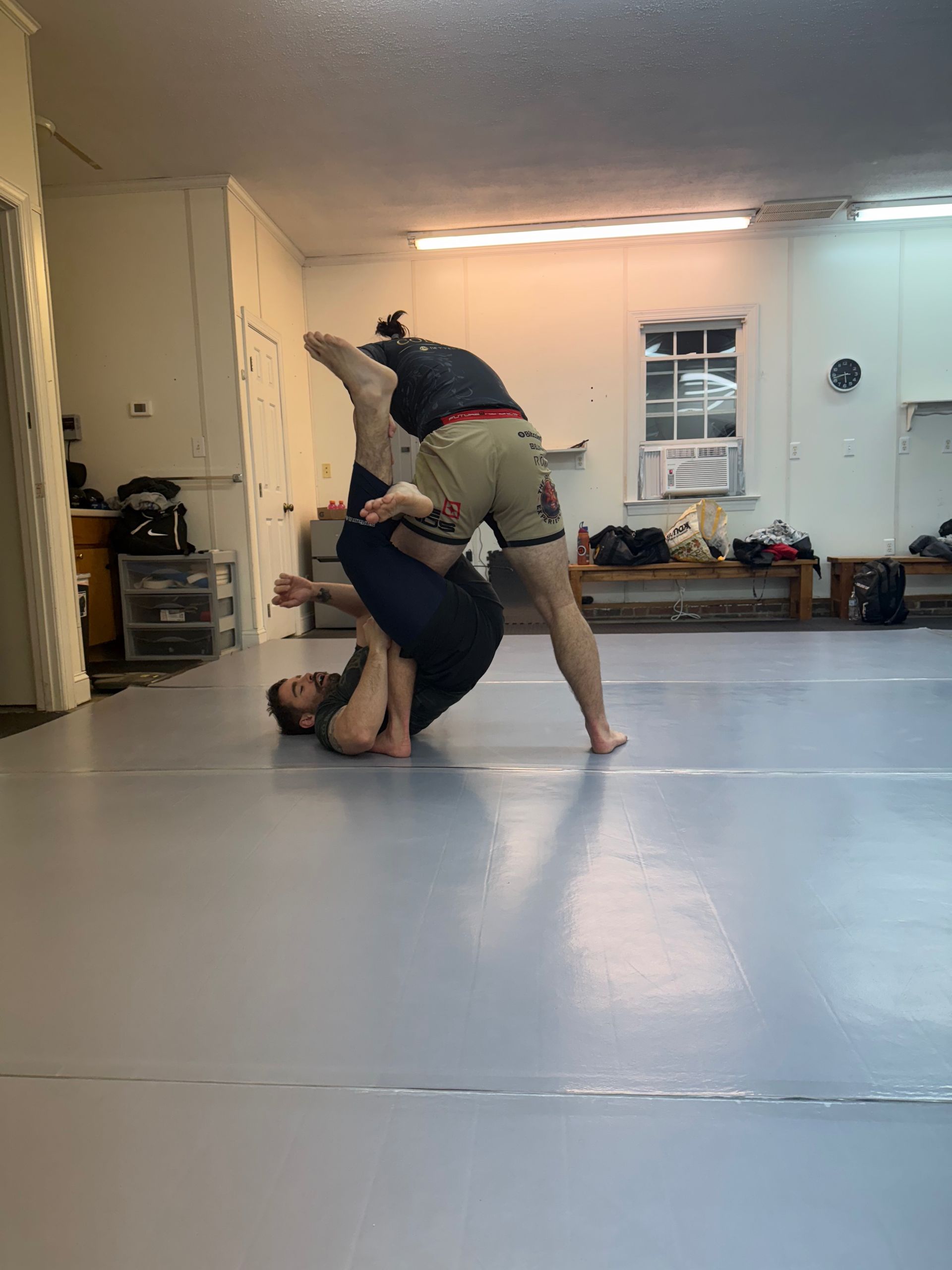 Two people practicing grappling on mats in a bright gym, one lifting the other upside down
