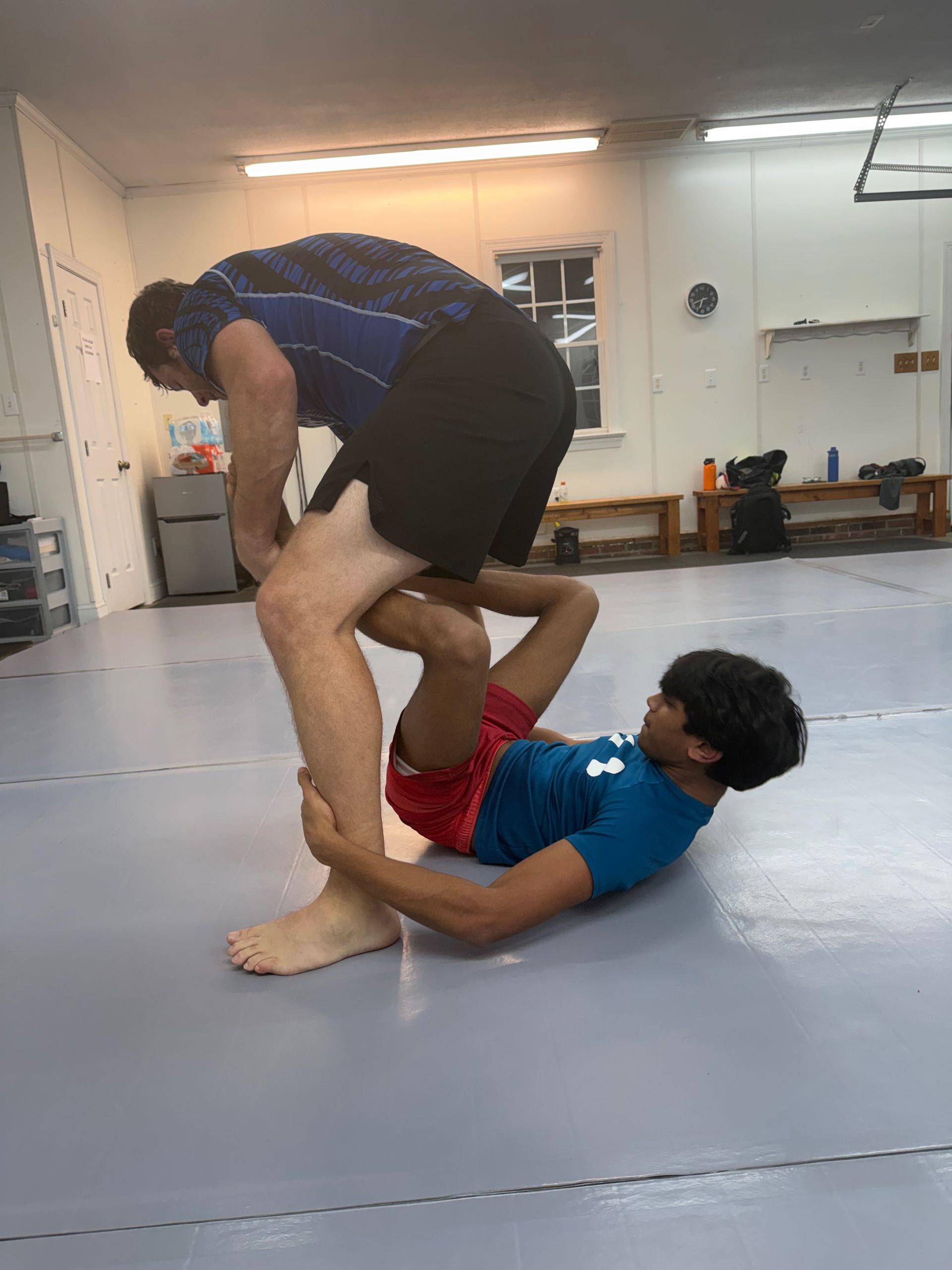 Two people practicing grappling on a mat in a gym, one standing over the other on the floor.