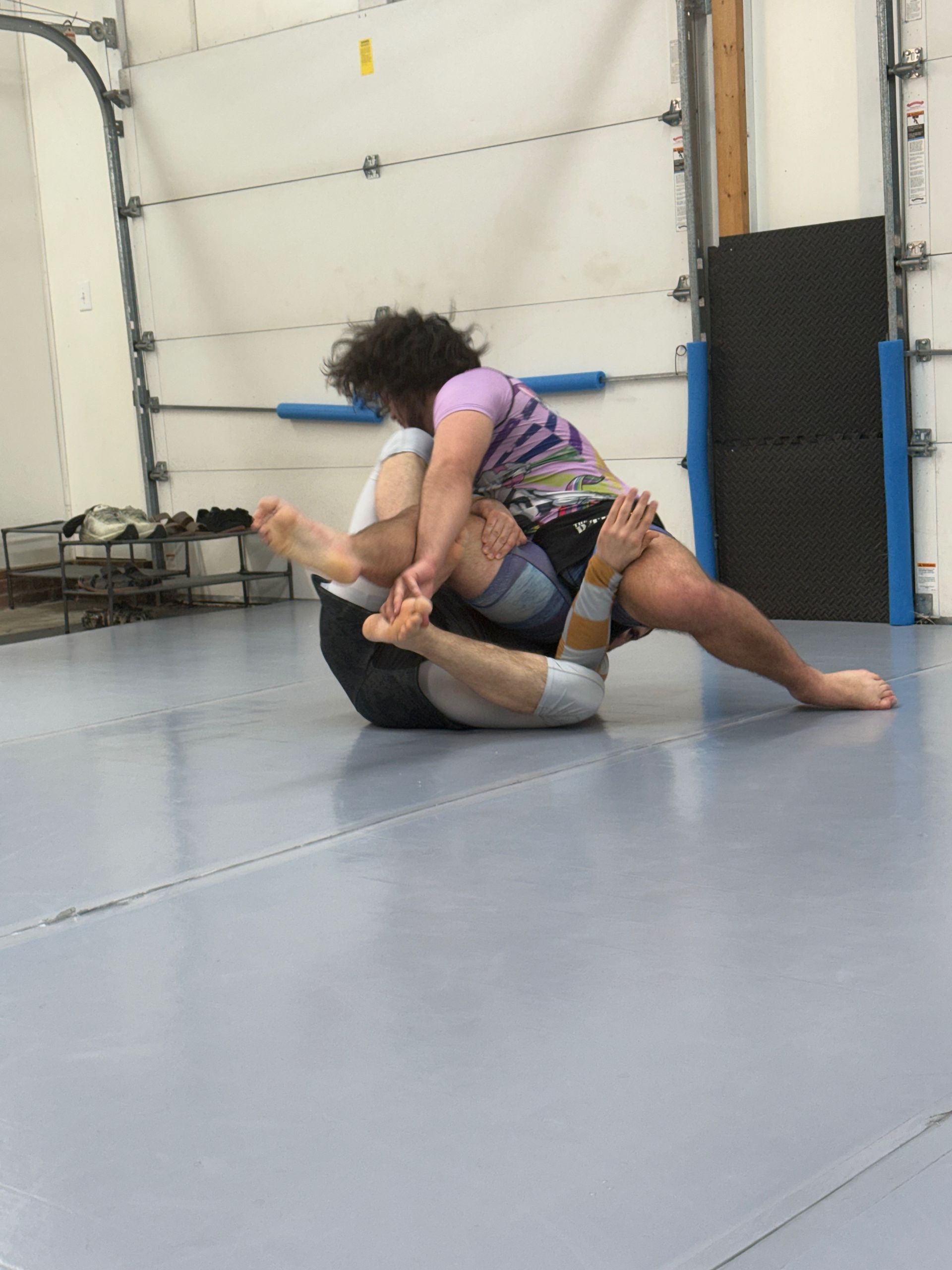 Two people grappling on a gym mat, one in a purple shirt on top in a grappling hold.