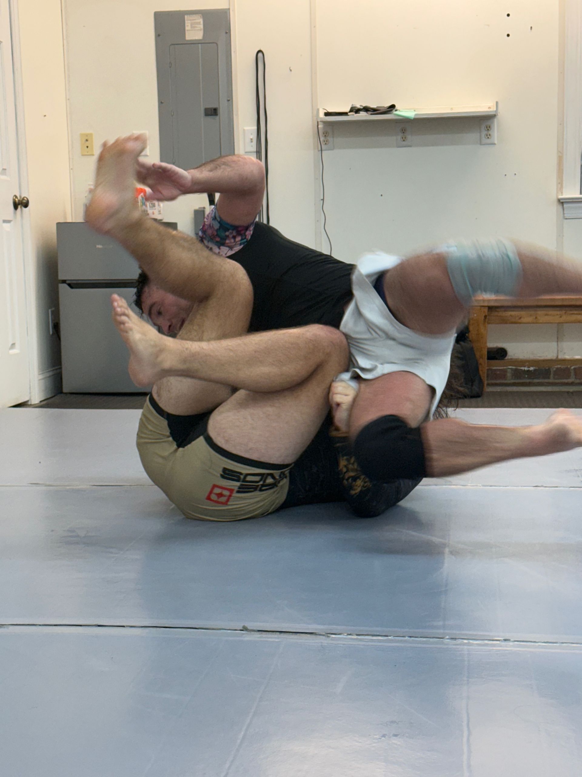 Two people grappling on a mat in a gym, one in a black shirt and one in a white shirt.