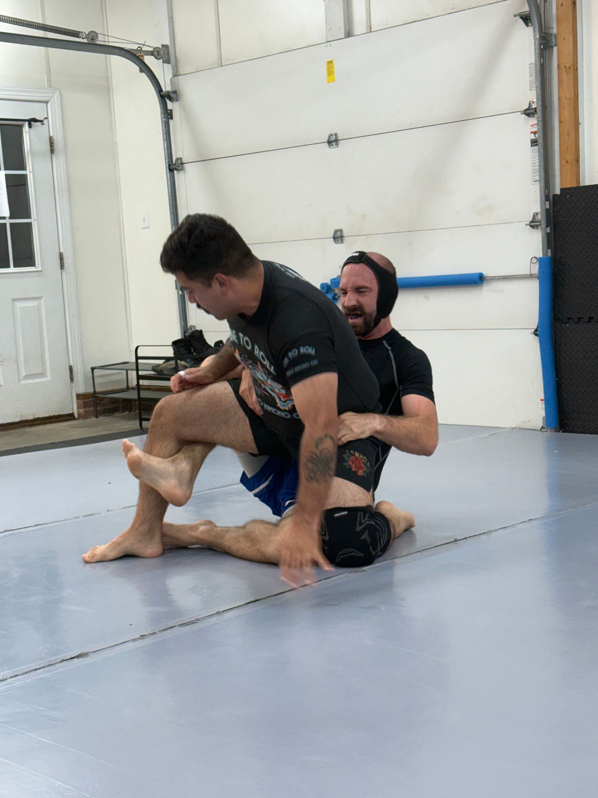Two men grappling on gym mats, one seated behind the other in a training hold.