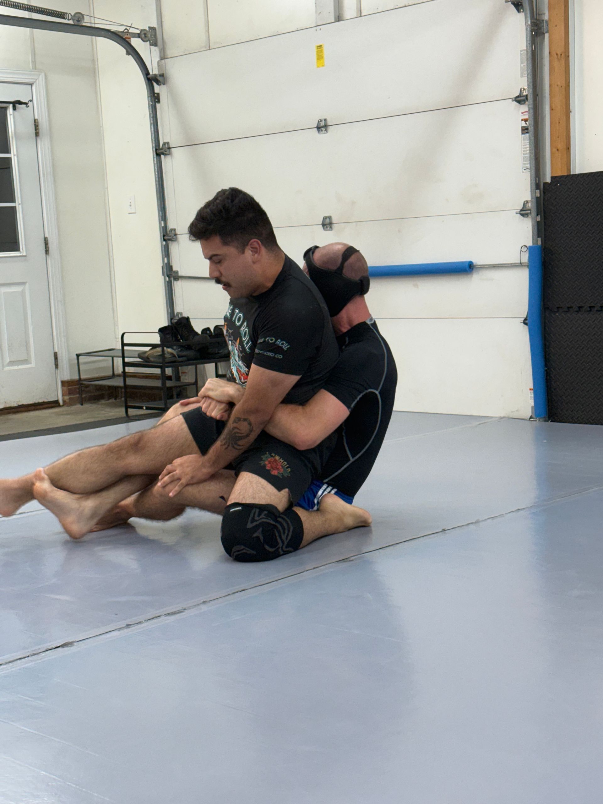 Two martial artists grappling on mats in a training gym, one seated and controlling the other from behind.