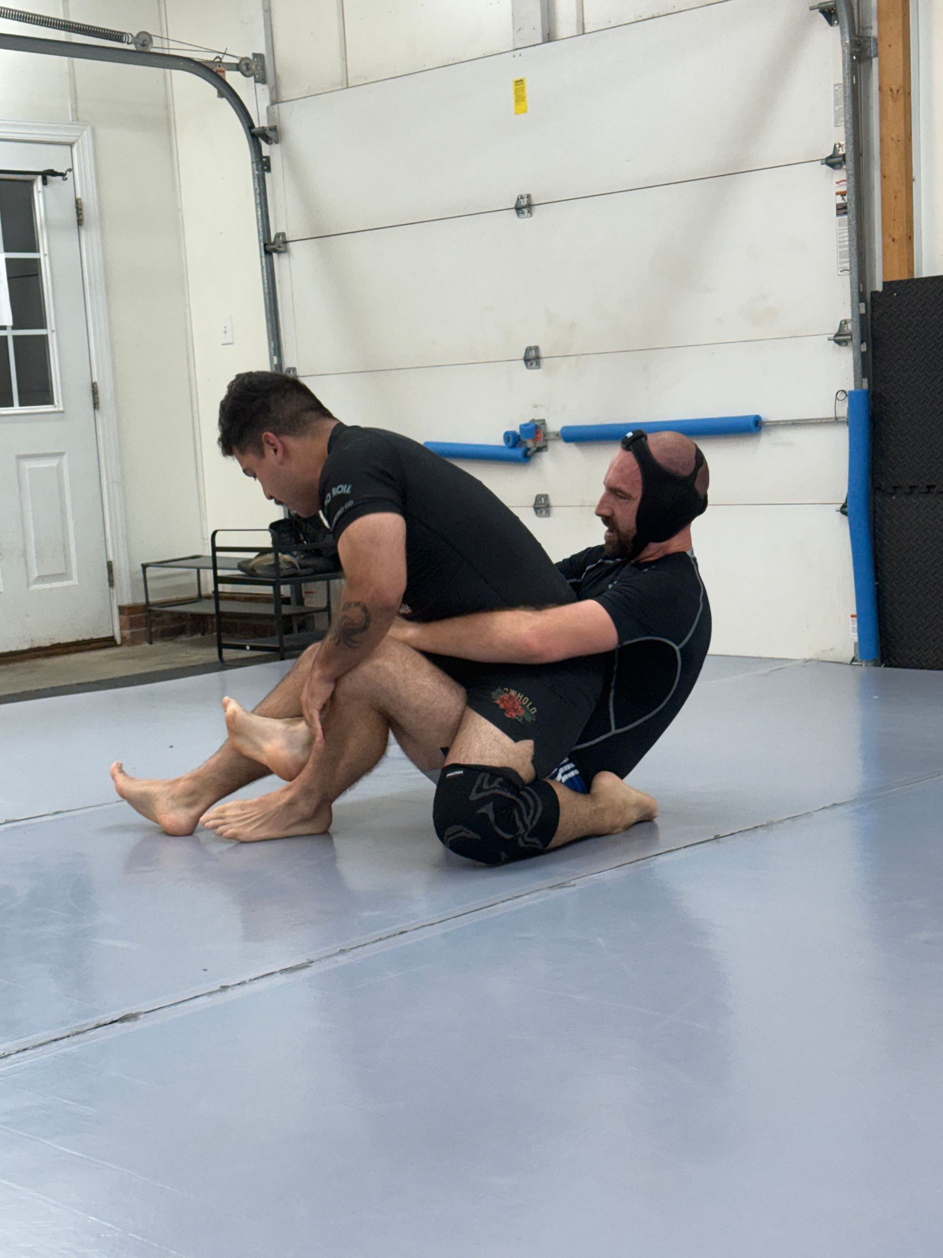 Two men grappling on mats in a gym, one seated and one kneeling in front of him.