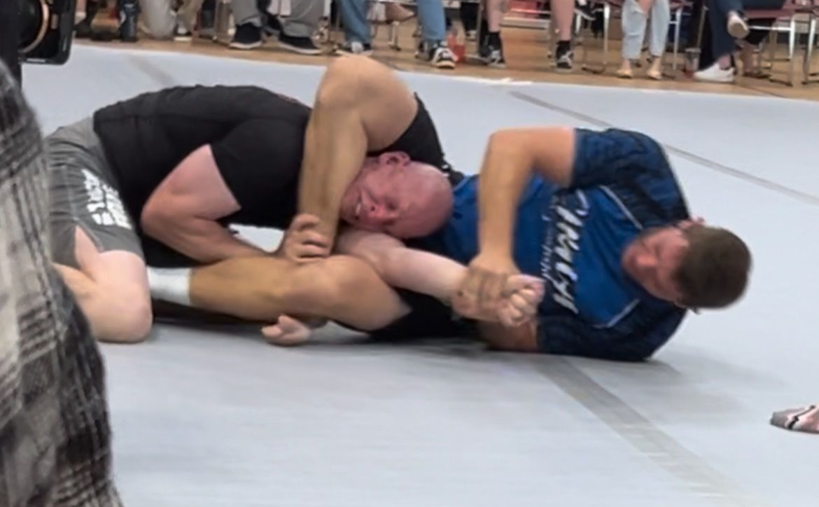 Two wrestlers grapple on a mat during a match, one in black and one in blue, with spectators in the background.