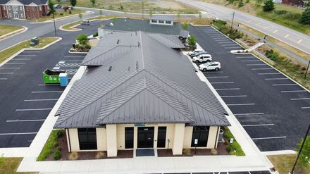 An aerial view of a tan commercial building with a dark, textured roof surrounded by parking spaces and a road.