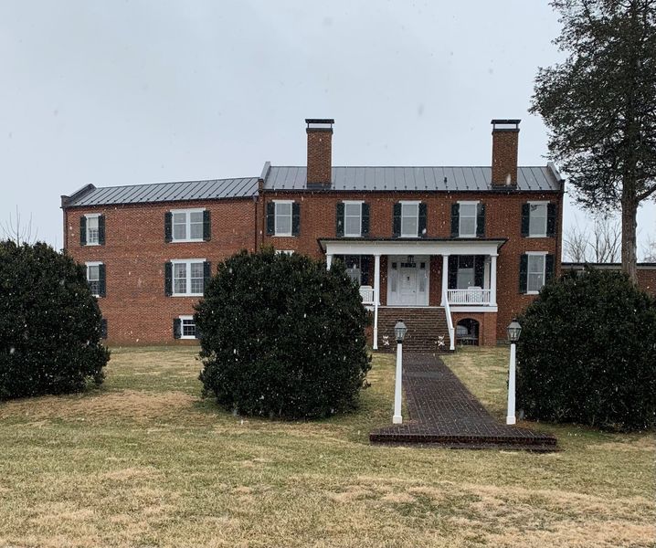 Red brick historic building with snow flurries, black shutters, and a walkway leading to a porch with white columns.
