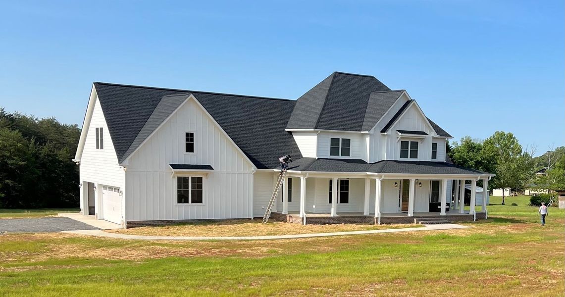 White farmhouse with a black roof, front porch, and garage, set in a grassy field under a blue sky.