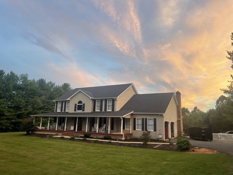 Two-story beige house with a dark roof and porch under a colorful sunset sky.